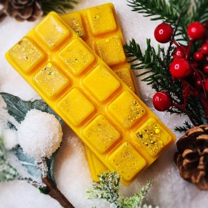 May include: Two yellow wax melt bars with glitter, arranged diagonally. Each bar has square sections. The background features pine needles, red berries, and a pine cone, creating a festive, seasonal display.