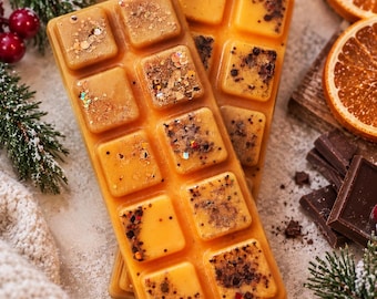 Puede incluir: Dos barras rectangulares de cera aromática en un tono naranja cálido, divididas cada una en diez segmentos cuadrados. La superficie de la cera está incrustada con varios elementos decorativos, incluyendo purpurina dorada y partículas botánicas oscuras.
