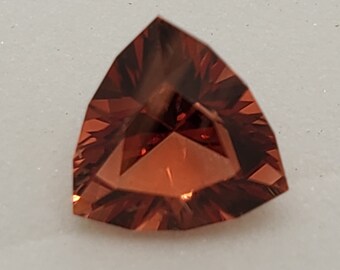 May include: A triangular, faceted gemstone with a reddish-brown color. The gem has a star-like pattern in the center.