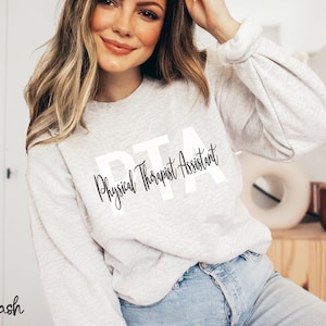 May include: A light gray crewneck sweatshirt featuring the large white block letters PTA with the words Physical Therapist Assistant written in a black cursive script font layered over the letters. The sweatshirt has long sleeves with ribbed cuffs.