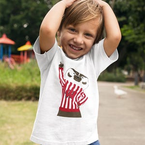 May include: A young child wearing a white t-shirt with a red and white striped badger mascot graphic. The badger is wearing a red and white striped shirt with a white letter "W" on the front. The child is smiling and has their hands on their head.