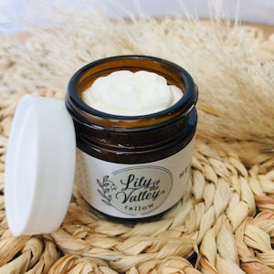 May include: A small glass jar with a white lid filled with a white cream. The jar has a label that says "Lily of the Valley Tallow" with a small floral design.