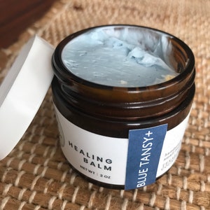 Könnte beinhalten: Ein kleines Glasgefäß mit einem blauen Deckel, gefüllt mit einem hellblauen Balsam. Das Etikett trägt die Aufschrift "Healing Balm" und "Blue Tansy+" in schwarzer Schrift auf weißem Hintergrund. Das Gefäß steht auf einer gewebten Oberfläche.
