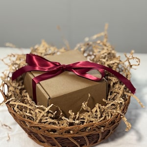 May include: A small, brown cardboard box tied with a burgundy satin ribbon, nestled in a woven wicker basket filled with shredded paper. The basket is brown and sits on a white surface, creating a gift presentation.