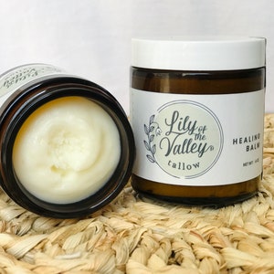 May include: Two amber glass jars with white lids containing a white balm. The jar on the right has a white label with the text "Lily of the Valley tallow" and "HEALING BALM NET WT 1.4 OZ".