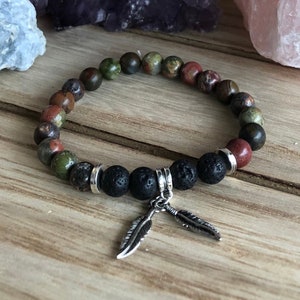 May include: A beaded bracelet with a mix of green, red, and brown stones. The bracelet has a black lava stone charm with two silver feather charms.