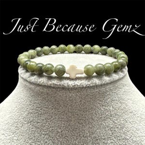 May include: Green beaded bracelet with a white cross-shaped charm. The bracelet is displayed on a gray surface against a black background. The text "Just Because Gemz" is written in white script above the bracelet.
