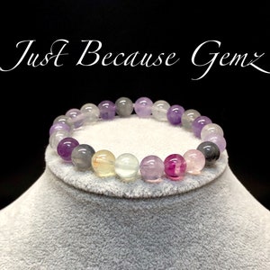Puede incluir: Una pulsera de cuentas con una variedad de piedras preciosas coloridas, que incluyen cuentas moradas, grises, rosas y transparentes. La pulsera se muestra sobre una superficie gris sobre un fondo negro. El texto de la imagen dice "Just Because Gemz".