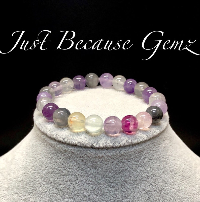 Puede incluir: Una pulsera de cuentas con una variedad de piedras preciosas coloridas, que incluyen cuentas moradas, grises, rosas y transparentes. La pulsera se muestra sobre una superficie gris sobre un fondo negro. El texto de la imagen dice "Just Because Gemz".