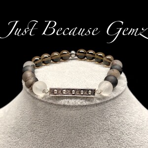 May include: A brown, gray, and white beaded bracelet with a silver bar charm. The bracelet has the text "Just Because Gemz" on the background.