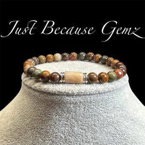 Puede incluir: Un brazalete de piedras preciosas marrones, verdes y rojas con un acento de cuentas de madera marrón claro. El brazalete está en un expositor gris.
