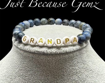May include: A beaded bracelet with blue and gray stones and white letter beads spelling "GRANDPA". The bracelet is displayed on a gray surface. The text "Just Because Gemz" is above the bracelet.