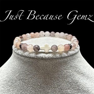 May include: A beaded bracelet with a white cross charm. The bracelet features a mix of pink, gray, and clear beads. The text "Just Because Gemz" is in the background.