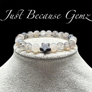 May include: A gray and black beaded bracelet with a black star charm. The bracelet is on a white display stand. "Just Because Gemz" is written in black text at the top of the image.