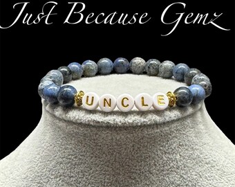 May include: A beaded bracelet with blue and gray stone beads. The bracelet has white letter beads that spell "UNCLE" with gold accents. The text "Just Because Gemz" is in the background.