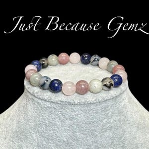 May include: A bracelet made of pink, blue, and gray gemstone beads. The bracelet is on a gray display stand.