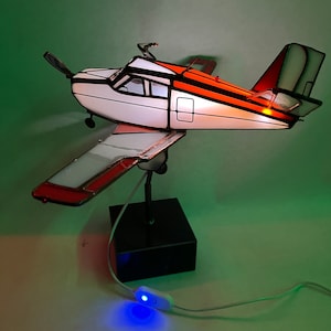 Puede incluir: L&aacute;mpara de avi&oacute;n de vidrieras con fuselaje blanco y naranja, h&eacute;lice negra y base negra. La l&aacute;mpara est&aacute; encendida y el interruptor est&aacute; encendido. Las alas y la cola tienen detalles en rojo. La l&aacute;mpara est&aacute; sobre una superficie blanca con fondo verde.