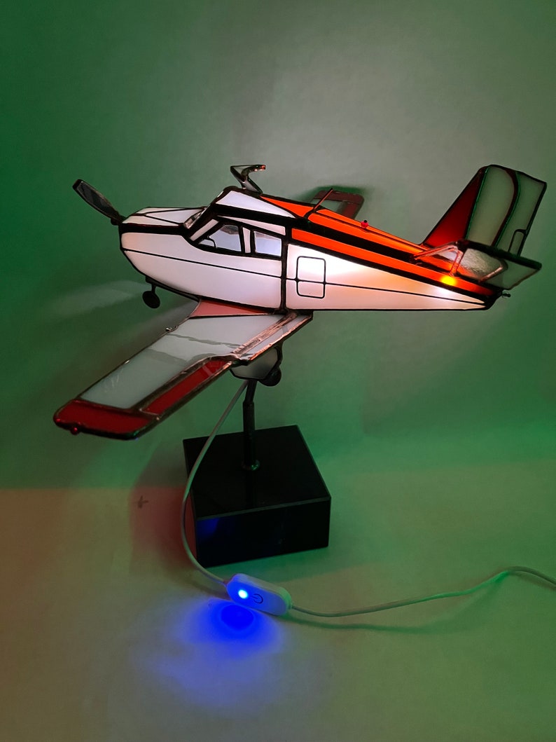 Puede incluir: L&aacute;mpara de avi&oacute;n de vidrieras con fuselaje blanco y naranja, h&eacute;lice negra y base negra. La l&aacute;mpara est&aacute; encendida y el interruptor est&aacute; encendido. Las alas y la cola tienen detalles en rojo. La l&aacute;mpara est&aacute; sobre una superficie blanca con fondo verde.