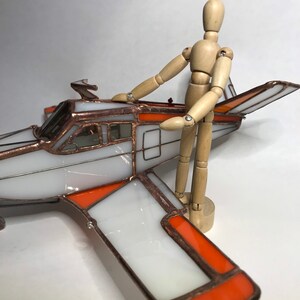 Puede incluir: Un modelo de avi&oacute;n de vidrieras con paneles blancos y naranjas, delineados en cobre. Una figura de madera posable est&aacute; de pie junto al avi&oacute;n. El avi&oacute;n tiene una h&eacute;lice y una luz roja en el ala.