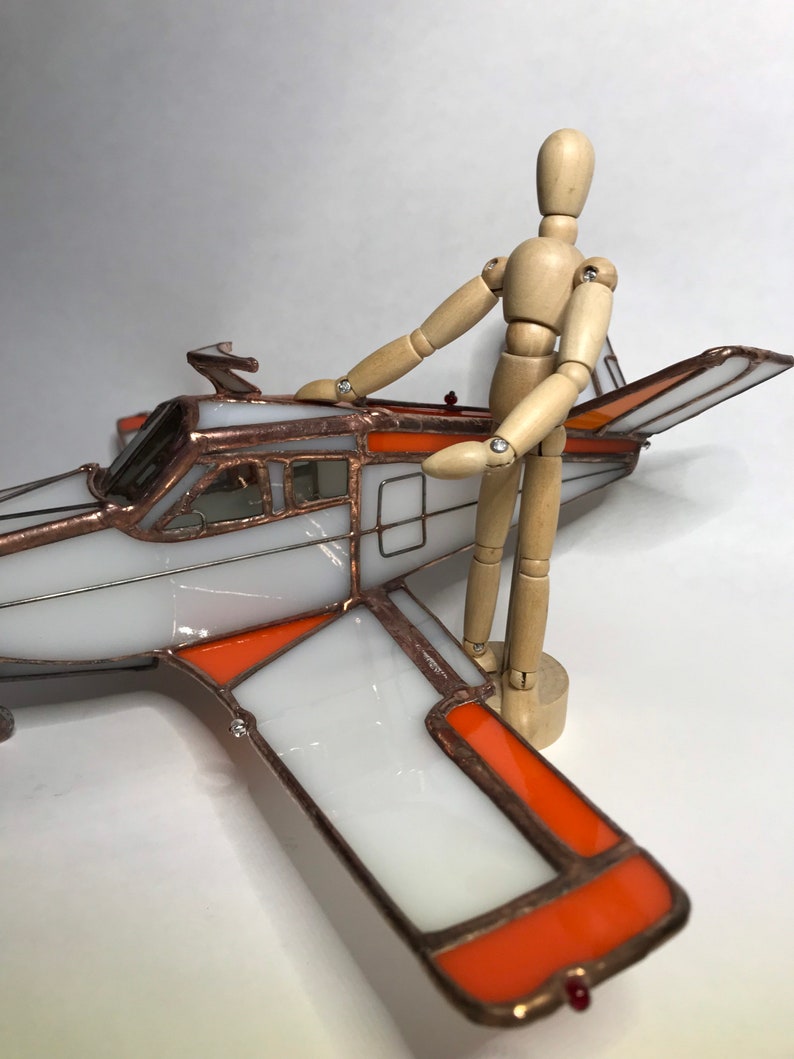 Puede incluir: Un modelo de avi&oacute;n de vidrieras con paneles blancos y naranjas, delineados en cobre. Una figura de madera posable est&aacute; de pie junto al avi&oacute;n. El avi&oacute;n tiene una h&eacute;lice y una luz roja en el ala.