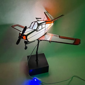 Puede incluir: Maqueta decorativa de avi&oacute;n con cuerpo blanco, ribetes negros y detalles en naranja. El modelo tiene h&eacute;lice, alas y cola. Est&aacute; montado sobre una base negra e iluminado con luz azul. El avi&oacute;n mide aproximadamente 30 cm de largo.