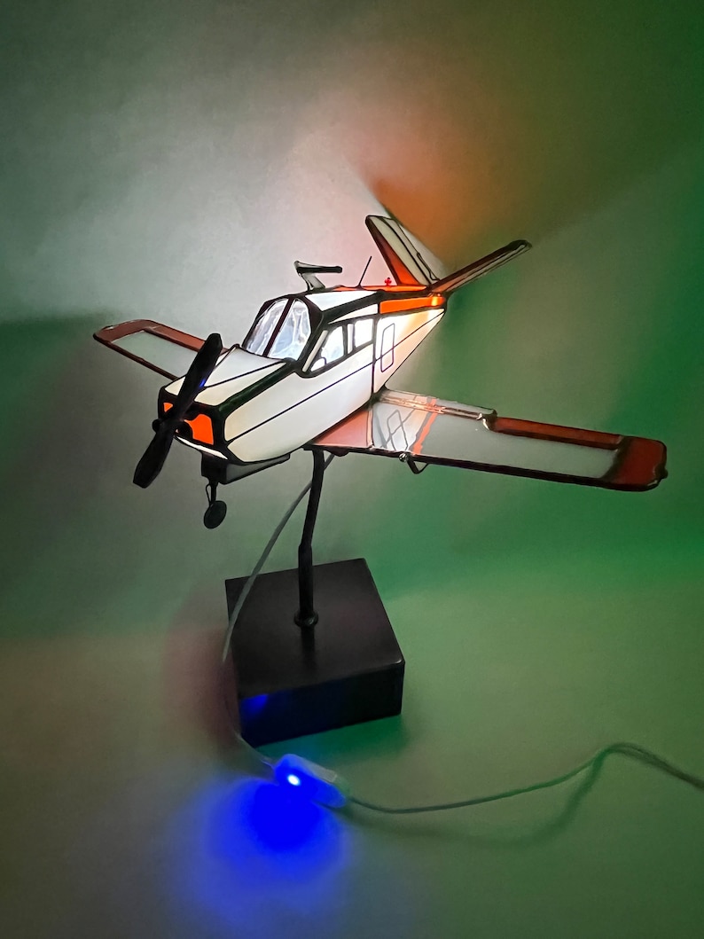 Puede incluir: Maqueta decorativa de avi&oacute;n con cuerpo blanco, ribetes negros y detalles en naranja. El modelo tiene h&eacute;lice, alas y cola. Est&aacute; montado sobre una base negra e iluminado con luz azul. El avi&oacute;n mide aproximadamente 30 cm de largo.
