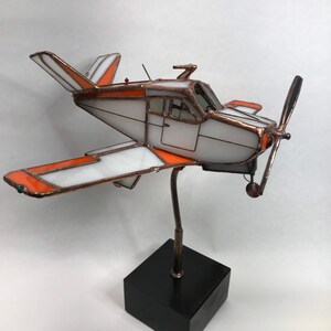 Puede incluir: Una escultura de avi&oacute;n de vidrieras con detalles en blanco y naranja, marco de cobre y una base de madera negra. La h&eacute;lice es negra y las alas tienen un dise&ntilde;o distintivo. La escultura est&aacute; montada sobre un soporte met&aacute;lico.