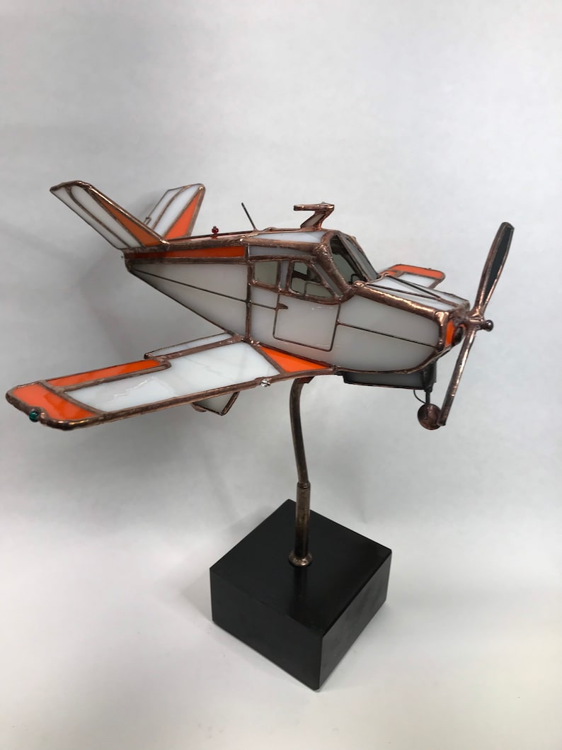 Puede incluir: Una escultura de avi&oacute;n de vidrieras con detalles en blanco y naranja, marco de cobre y una base de madera negra. La h&eacute;lice es negra y las alas tienen un dise&ntilde;o distintivo. La escultura est&aacute; montada sobre un soporte met&aacute;lico.