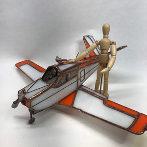 Puede incluir: Un modelo de avi&oacute;n de vidrieras con detalles en blanco y naranja. Las alas y la cola tienen paneles blancos con bordes naranjas. El cuerpo del avi&oacute;n es plateado con un marco color cobre. Una figura de madera articulada est&aacute; de pie junto al avi&oacute;n.