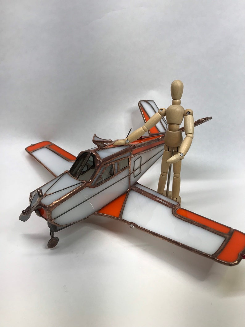 Puede incluir: Un modelo de avi&oacute;n de vidrieras con detalles en blanco y naranja. Las alas y la cola tienen paneles blancos con bordes naranjas. El cuerpo del avi&oacute;n es plateado con un marco color cobre. Una figura de madera articulada est&aacute; de pie junto al avi&oacute;n.