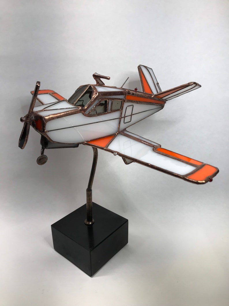 Puede incluir: Una escultura de avi&oacute;n de vidrieras con detalles en blanco y naranja, montada sobre una base cuadrada negra. El modelo de avi&oacute;n presenta una h&eacute;lice, alas y una cabina detallada. El marco de metal a&ntilde;ade una est&eacute;tica vintage.