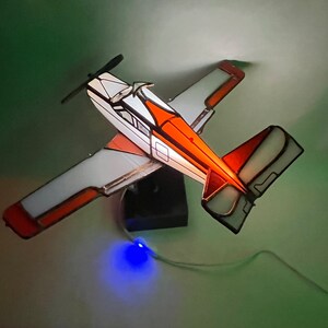 Puede incluir: Maqueta de avi&oacute;n de vidrieras con detalles en blanco, naranja y negro. Las alas y la cola son blancas con ribetes naranjas. El fuselaje es blanco con detalles en naranja y negro. Se ve una h&eacute;lice y una base negras.