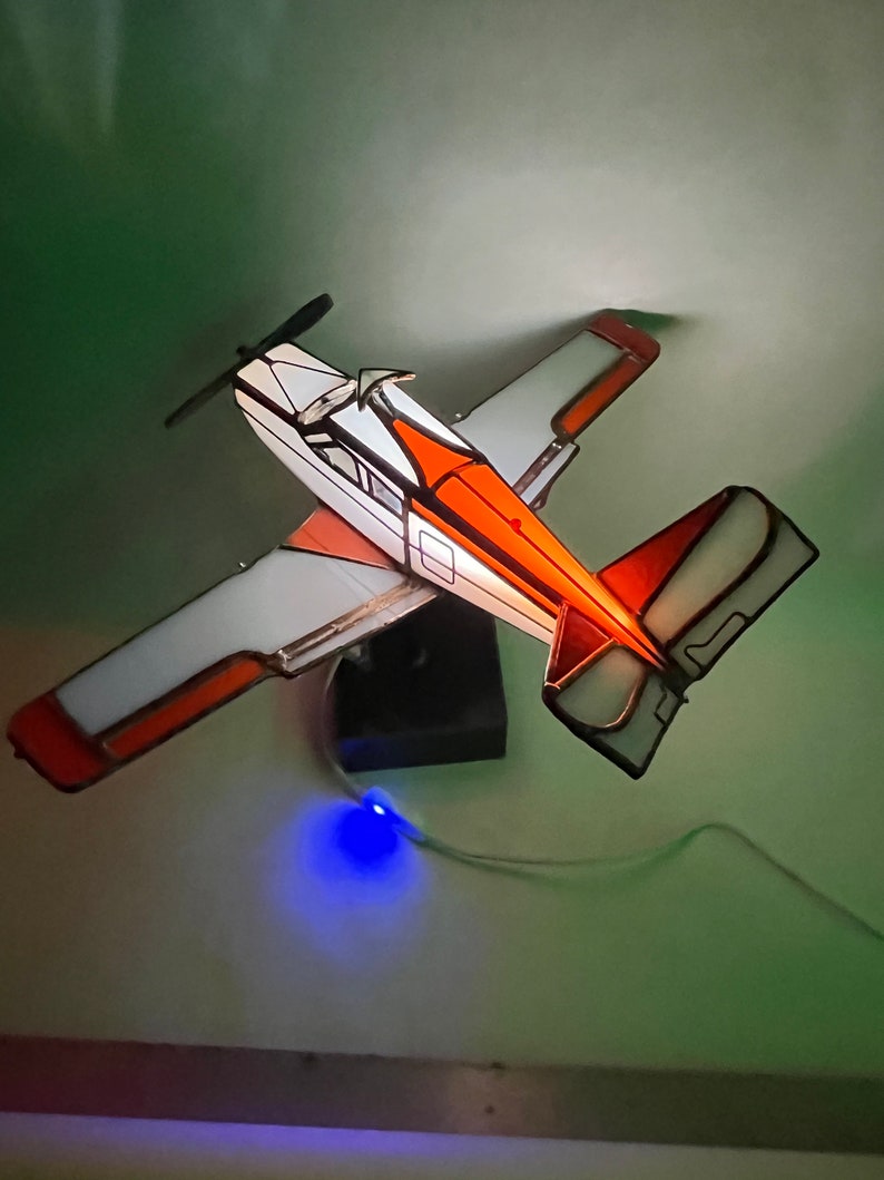 Puede incluir: Maqueta de avi&oacute;n de vidrieras con detalles en blanco, naranja y negro. Las alas y la cola son blancas con ribetes naranjas. El fuselaje es blanco con detalles en naranja y negro. Se ve una h&eacute;lice y una base negras.