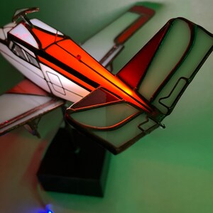 Puede incluir: L&aacute;mpara de avi&oacute;n de vidrieras con paneles naranjas, blancos y rojos. La l&aacute;mpara tiene una base negra y est&aacute; iluminada con una luz azul. Las alas y la cola est&aacute;n detalladas con l&iacute;neas de plomo.