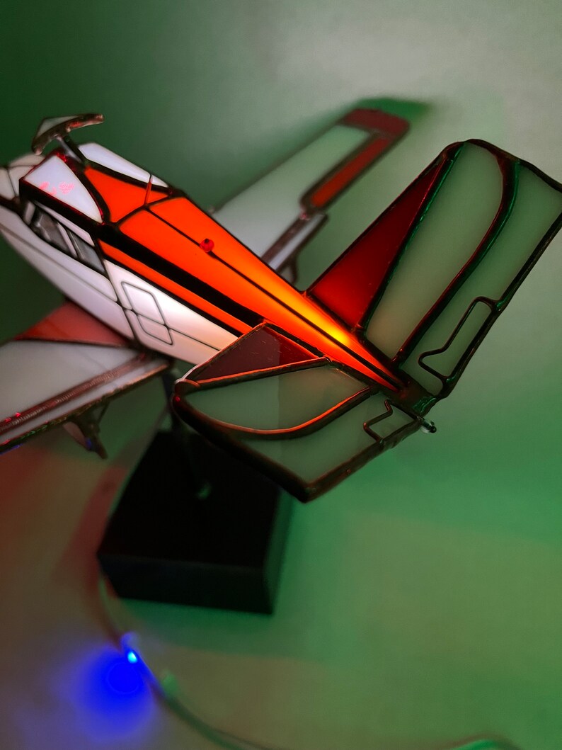 Puede incluir: L&aacute;mpara de avi&oacute;n de vidrieras con paneles naranjas, blancos y rojos. La l&aacute;mpara tiene una base negra y est&aacute; iluminada con una luz azul. Las alas y la cola est&aacute;n detalladas con l&iacute;neas de plomo.