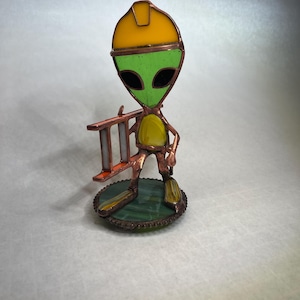 Peut inclure: Figurine en vitrail représentant un extraterrestre portant un casque jaune et tenant une petite échelle. L'extraterrestre a un visage vert, des yeux noirs et un corps jaune. La figurine est montée sur une base circulaire verte et marron.