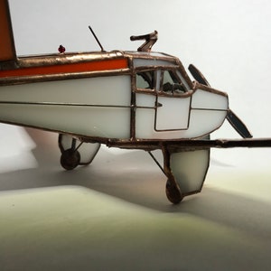Puede incluir: Maqueta de avi&oacute;n de vidrieras hecha a mano. El avi&oacute;n tiene un cuerpo blanco con detalles naranjas en las alas y la parte superior. El modelo tiene una h&eacute;lice, ruedas y una peque&ntilde;a luz roja. El marco es de cobre.