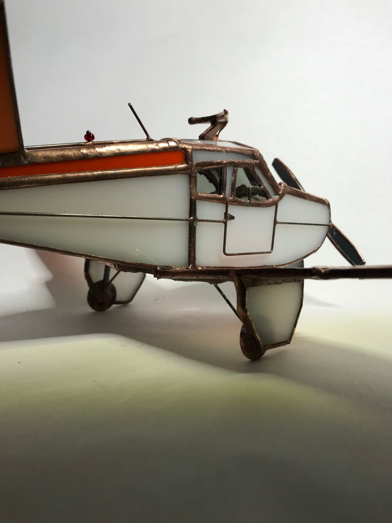 Puede incluir: Maqueta de avi&oacute;n de vidrieras hecha a mano. El avi&oacute;n tiene un cuerpo blanco con detalles naranjas en las alas y la parte superior. El modelo tiene una h&eacute;lice, ruedas y una peque&ntilde;a luz roja. El marco es de cobre.