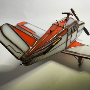 Puede incluir: Maqueta de avi&oacute;n artesanal de vidrieras. El modelo presenta paneles blancos y naranjas con l&iacute;neas de soldadura color cobre. La h&eacute;lice y otros detalles tambi&eacute;n son de color cobre. El avi&oacute;n tiene un dise&ntilde;o cl&aacute;sico y vintage.