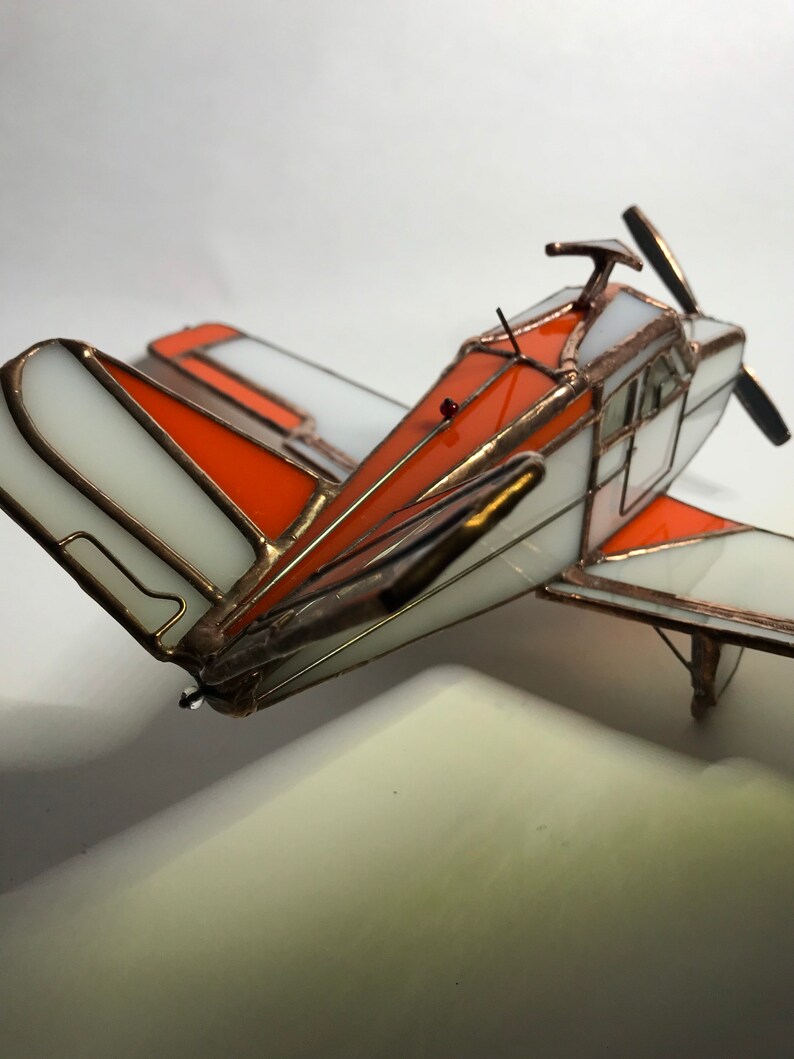 Puede incluir: Maqueta de avi&oacute;n artesanal de vidrieras. El modelo presenta paneles blancos y naranjas con l&iacute;neas de soldadura color cobre. La h&eacute;lice y otros detalles tambi&eacute;n son de color cobre. El avi&oacute;n tiene un dise&ntilde;o cl&aacute;sico y vintage.