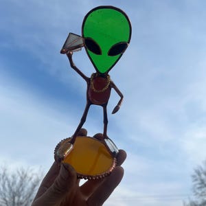 May include: Stained glass alien figure with a green head, black eyes, and a gold chain. The alien holds a small pyramid and stands on a yellow and gold base. The figure is set against a blue sky.