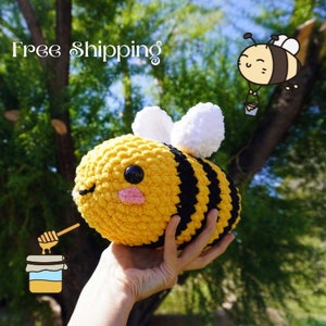 May include: A crocheted bee plush toy with black and yellow stripes, white wings, and a cute face. The bee is being held by a hand in front of a green leafy background.