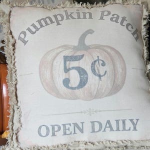 May include: A square off-white throw pillow with frayed edges featuring a printed graphic of a pumpkin. The text reads Pumpkin Patch, 5 cents, and OPEN DAILY in a grey font. The design is centered on the front of the pillow fabric.