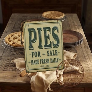 May include: A rustic wooden table displays a vintage metal sign that reads "PIES FOR SALE - MADE FRESH DAILY." Two pies sit on the table, one on a wire rack. A linen cloth and rolling pin are also present, suggesting a bakery or kitchen setting.