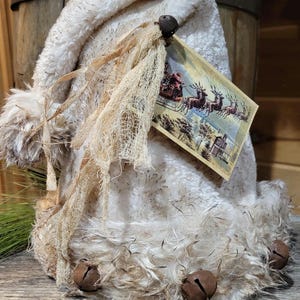 May include: A white faux fur Santa hat with a vintage postcard attached. The postcard features a scene of Santa Claus flying in a sleigh pulled by reindeer. The hat has a frayed edge and is decorated with bells.