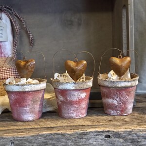 May include: Three small, rustic red and white painted buckets with metal heart ornaments hanging from wire handles. The buckets are filled with white paper doilies, and sit on a wooden surface.