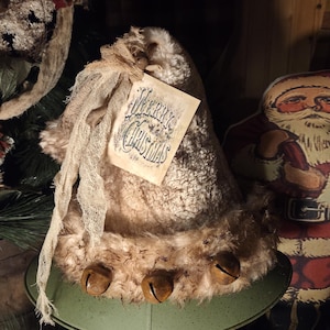 May include: A rustic Christmas hat with a tag that reads "Merry Christmas." The hat is beige with a furry texture and adorned with three bronze bells. A beige fabric ribbon is attached to the hat.