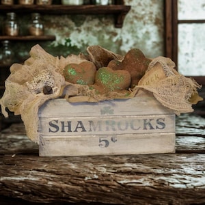 May include: A rustic wooden crate with the word "SHAMROCKS" printed on the front, containing heart-shaped decorations with glitter accents. The crate is filled with burlap and decorative elements, evoking a vintage aesthetic.