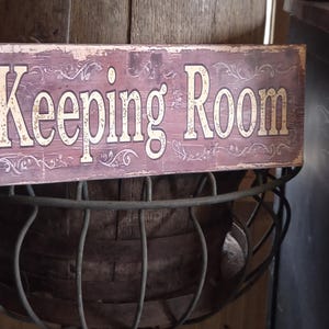 May include: A rectangular wooden sign with the words "Keeping Room" in large, off-white letters. The sign has a distressed, vintage look with a faded burgundy background and decorative floral accents. It is mounted above a wire basket.
