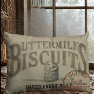 May include: Rectangular pillow with the words "BUTTERMILK BISCUITS" in large, distressed lettering. Below, it reads "BAKED FRESH DAILY." A graphic of a biscuit and a circular stamp with a farm scene are also visible. The pillow is beige.
